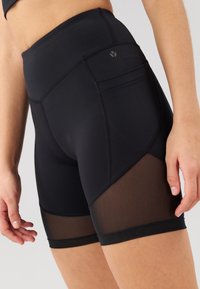 Sorte idrettsshorts med en tettsittende design, med meshpaneler langs sidene. Laget av elastisk stoff med synlige sømmer.