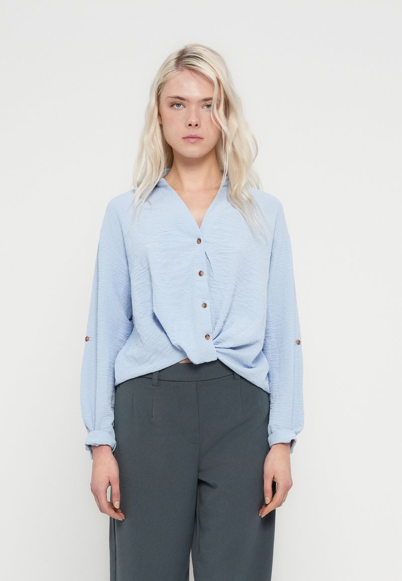 Mulher loira vestindo uma blusa azul clara texturizada com botões, enfiada numa calça cinza escura de cintura alta, em pé contra um fundo branco.