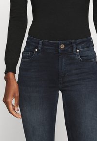 Jeans de denim azul-escuro com cintura média, bolsos frontais e fecho em botão de metal. Textura suave, desbotamento mínimo, corte reto.