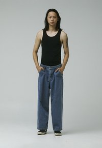 Black sleeveless tank top, loose blue denim jeans with a faded texture, and dark sneakers. Subject has long hair and a tattoo on the arm.