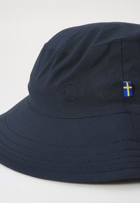 Námornícky modrý vedierkový klobúk vyrobený z jemnej textílie, s decentne embosovaným logom líšky a malou švédskou vlajkou na boku.