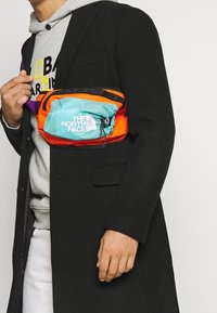 La borsa a tracolla The North Face presenta un corpo turchese con accenti arancioni, una tasca in rete e un logo bianco ben visibile. Cinghia regolabile viola.