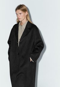 Cappotto di lana nera con vestibilità rilassata, design a un solo petto, grandi risvolti e tasche laterali. Indossato sopra una camicia di colore chiaro.