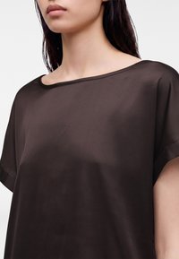 Femme portant un haut court à manches courtes en soie brun foncé avec un large col rond, montrée des épaules à la partie supérieure de la poitrine.