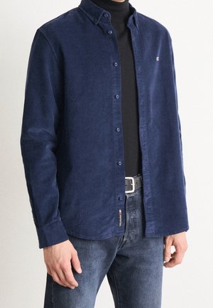 Mand iført mørkeblå kordfløjlskjorte med knapper over en sort højhalset trøje, kombineret med blå jeans og sort bælte.