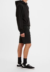Sweat à capuche noir avec une poche avant, associé à un short noir. Porté avec des chaussettes noires et des baskets basses noires avec des accents blancs.