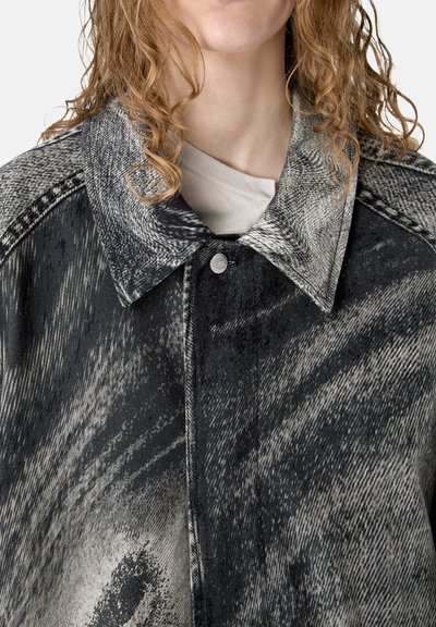 Nahaufnahme einer Person, die einen schwarz-grau gemusterten, geknöpften Mantel mit weitem Kragen trägt und lockiges, hellbraunes Haar hat.