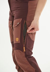 Herrarnas utomhusbyxor i mörkröd och brun tyg. Har sidofickor med dragkedja, meshfoder och läderdetaljer.