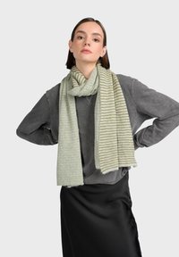 Ljusgrön randig scarf i mjukt, texturerat material, virad runt halsen, bärs med en grå tröja och svart kjol.