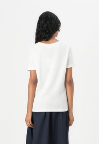 Weißes Baumwoll-T-Shirt mit rundem Ausschnitt und kurzen Ärmeln, das eine glatte Textur und eine figurbetonte Form aufweist. Sichtbare Rückennähte.