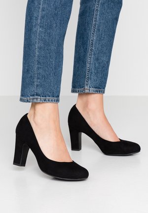 Pieds portant des escarpins en daim noir à talons hauts associés à un jean bleu coupé au-dessus des chevilles sur un sol clair.