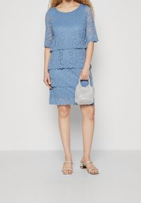 Robe en dentelle bleu clair avec un design superposé, manches courtes, bords festonnés. Accessoirisée avec un petit sac à main à motif et des sandales argentées.