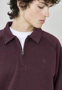 Pull à col zippé en tissu violet profond. Présente un logo subtil sur la poitrine et une texture lisse. Conçu pour un look ajusté.