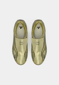 Zlati športni čevlji z elastiko za natik, črne poudarke, ukrivljeno obliko, gladko teksturo, z izrazitim logotipom in minimalističnim dizajnom.