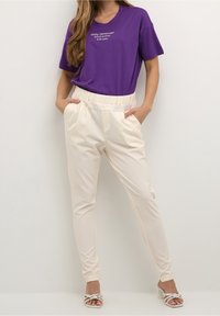 T-shirt violet à manches courtes avec texte blanc, associé à un pantalon fuselé de couleur crème avec taille élastique et poches latérales.