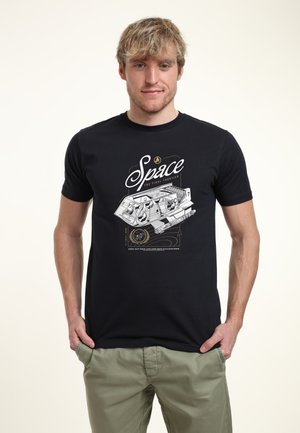 Ung mand med blondt hår iført sort t-shirt med hvidt rumskibsdesign og ordet "Space" over det, kombineret med grønne bukser.