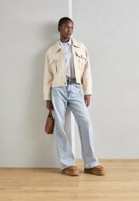 Giacca cropped beige con due tasche frontali, camicia a righe blu e bianca, jeans azzurri, scarpe platform marroni e borsa marrone.