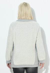 Persona con capelli biondi alla spalla che indossa un maglione grigio chiaro a maniche lunghe e pantaloni scuri, vista da dietro su uno sfondo bianco.