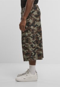 Bărbat purtând pantaloni scurți cargo camuflaj de lungime medie cu buzunare aplicate și adidași albi, stând în profil pe un fundal simplu.