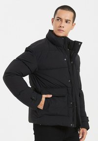 Doudoune noire avec un col montant, dotée d'une fermeture éclair, de boutons-pression et d'une poche avant. Texture lisse et design isolé.