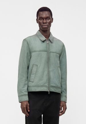 Jeune homme portant une veste verte zippée avec col et un pantalon noir, debout devant un fond clair uni.