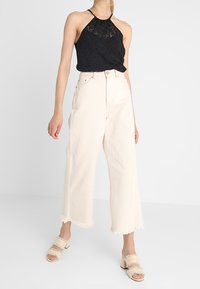 Black lace halter top paired with high-waisted, wide-leg, off-white denim pants with frayed hems and beige sandals with a textured strap.