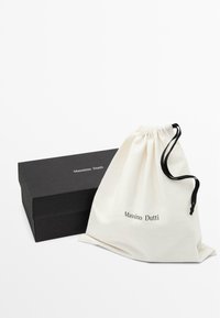 Cotton drawstring bag in cream with black cord, featuring "Massimo Dutti" in black text. Accompanied by a sleek black box.