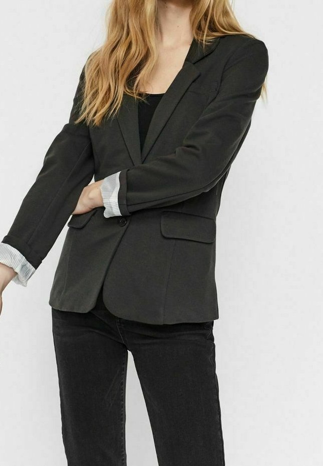 Femme portant un blazer noir aux manches retroussées sur un haut noir et un jean noir, debout devant un fond uni.