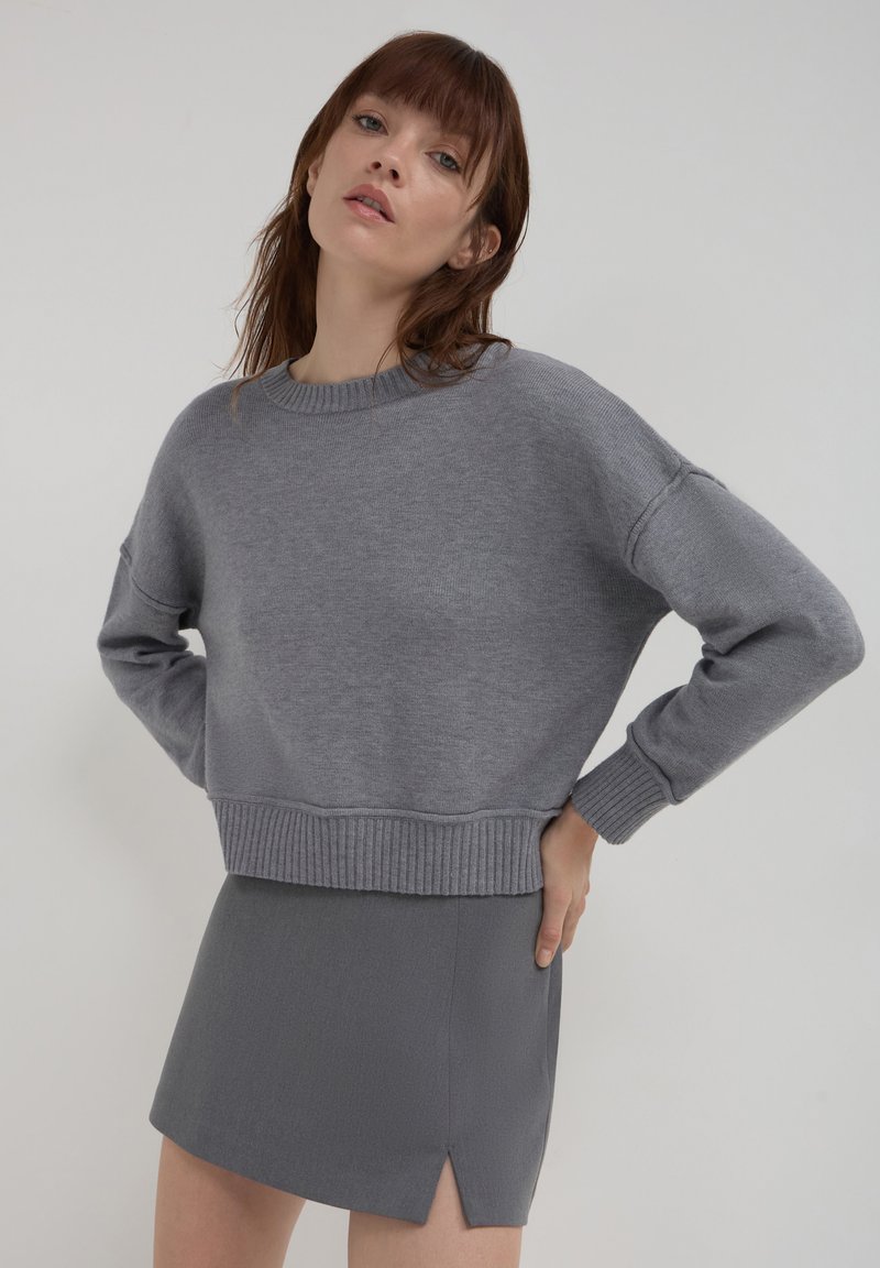 Pull gris court avec ourlet et poignets côtelés, présentant une coupe ample et un col rond. Associé à une jupe mini grise assortie avec une fente sur le côté.