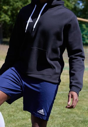 Osoba ubrana w czarną bluzę z kapturem Adidas i niebieskie spodenki Adidas, stojąca na trawie z lekko uniesionym jednym kolanem.