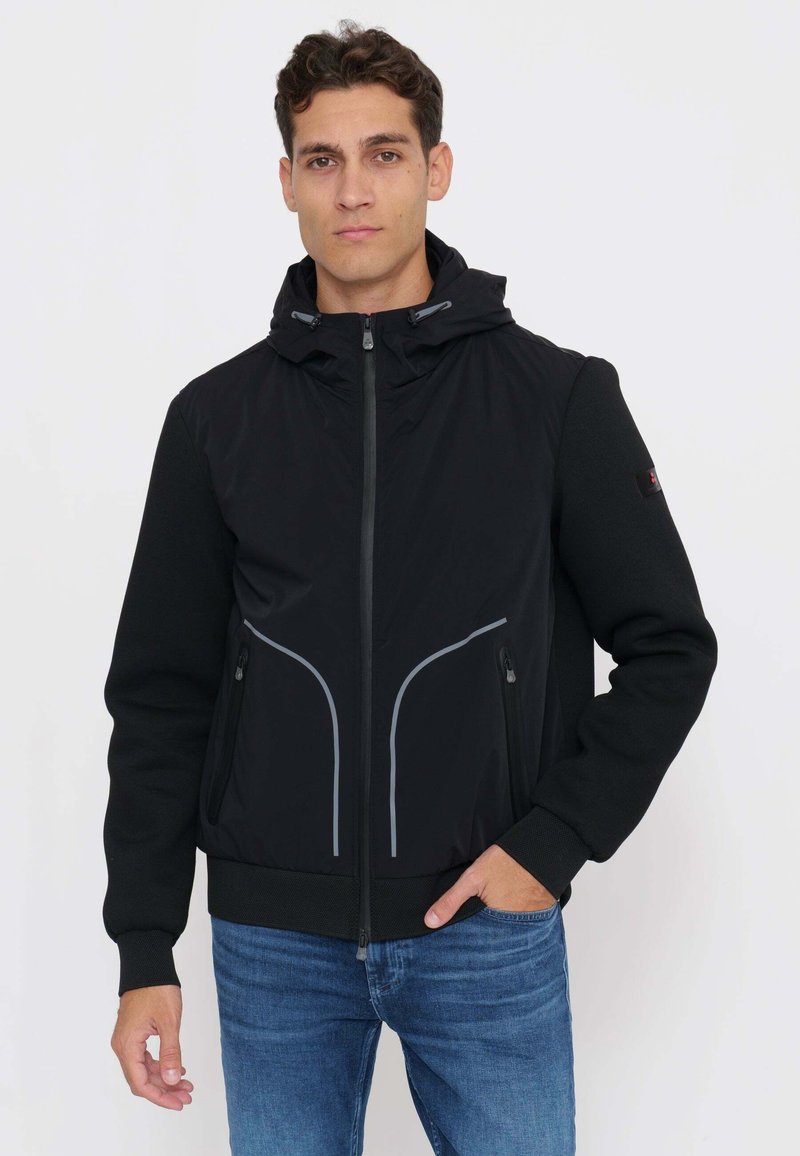 Uomo che indossa una giacca nera con cappuccio e piping riflettente, jeans blu, in piedi davanti a uno sfondo chiaro uniforme con una mano in tasca.