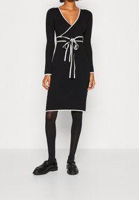 Robe noire côtelée à manches longues, col en V, bordure blanche et ceinture à la taille. Associée à des chaussures noires et des collants opaques.