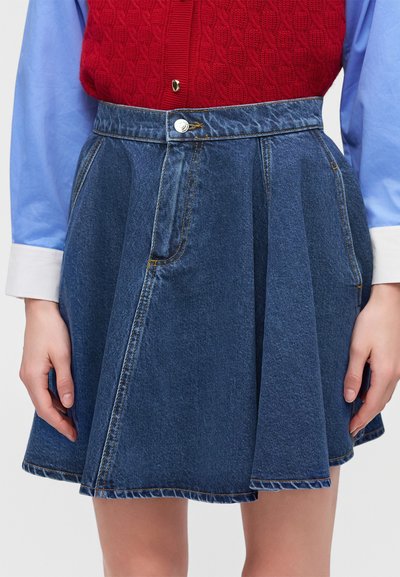 Personne portant une jupe plissée en denim bleu avec des poches avant, un gilet en tricot rouge et une chemise bleu clair avec des poignets blancs.