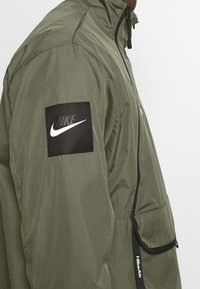 Chaqueta ligera de color verde oliva con un cuello alto, cierre de cremallera y parche de logo en la manga izquierda. Cuenta con un bolsillo con cremallera y detalles ajustables.