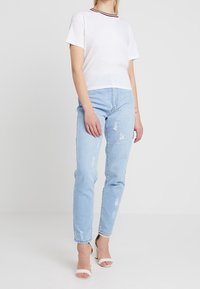 Bílá košile s krátkým rukávem a červeným a námořnickým pruhovaným výstřihem, v kombinaci se světle modrými džíny s efektem opotřebení. Obuty bílé sandály na podpatku.