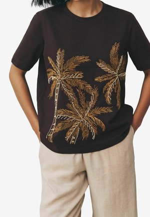 Person wearing a black short-sleeve shirt with gold and white palm tree designs, paired with light beige pants, hands in pockets.