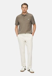 Braunes kurzärmeliges Poloshirt mit einer Tasche, kombiniert mit weißen Hosen und Sneakern mit braunen und schwarzen Akzenten.