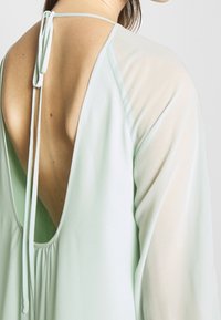Robe vert menthe clair avec un dos en V profond, des manches longues transparentes et une fermeture à nouer au niveau du cou. Texture lisse avec des détails froncés.