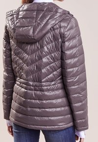 Vadderad, huvad jacka i en dämpad grå färg. Har ett chevron-mönster och ribbade fållar. Tillverkad av glänsande, vadderat material.