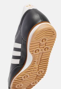 Black leather sneaker with white stripes, rubber gum sole, and orange accents. Features rounded toe and stitched details for durability.