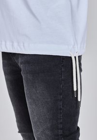 Bijela pamučna majica ravnog ruba s bočnim prorezima, uparena s tamnosivim trapericama s glatkom teksturom i kontrastnim vrpcama za vezanje.