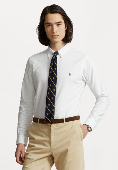 Vállig érő sötét hajú férfi fehér gombos ingben, tengerészkék csíkos nyakkendővel, bézs nadrággal és barna övvel, egyik kezét a zsebében tartva áll.