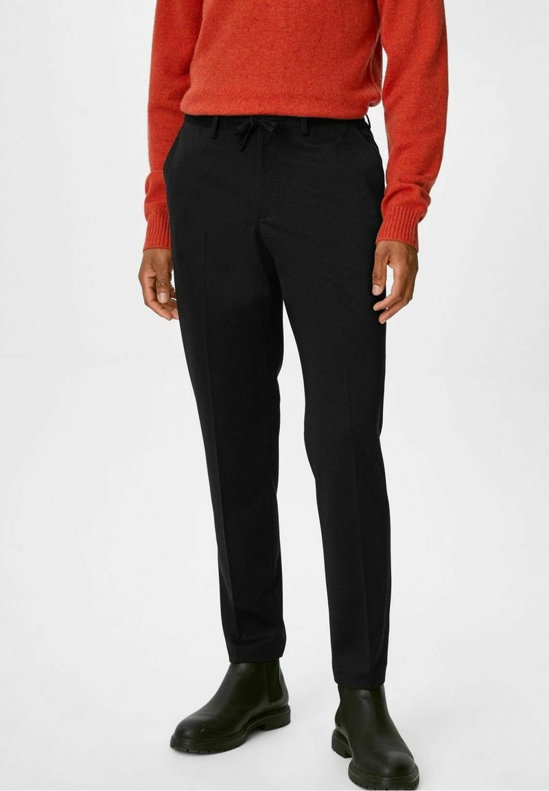 Svarta byxor med skräddarsydd passform, utrustade med sidfickor och dragsko i midjan. Stylade med en röd stickad tröja och svarta Chelsea-stövlar.