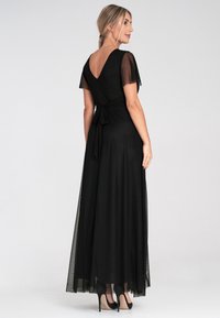Schwarzes Abendkleid mit transparenten Flatterärmeln, einem tiefen V-Ausschnitt hinten und einer gebundenen Taillendetail. Der Rock fließt bis zum Boden. Getragen mit schwarzen Absatzschuhen.