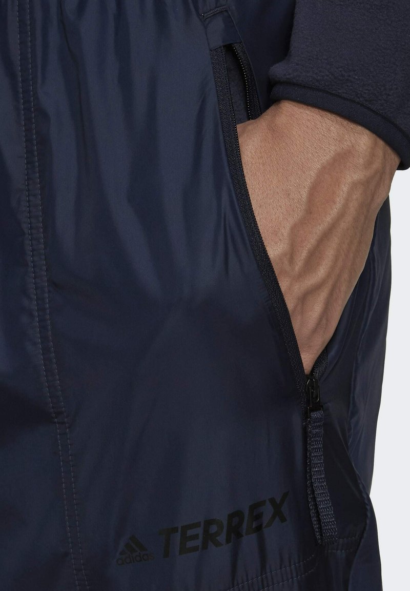 Chaqueta Terrex azul marino con tejido texturizado, bolsillos con cremallera y un logo prominente. Incluye un primer plano de una mano en el bolsillo.