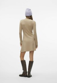 Abito a maglia beige con maniche lunghe, orlo svasato, indossato con stivali marroni fino al ginocchio e un berretto a maglia di un leggero viola. Texture liscia, design casual.