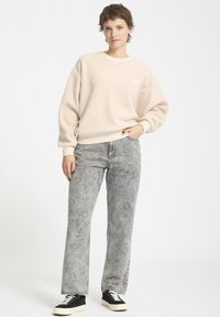 Jasno beżowy polarowy sweatshirt z ściągaczami, zestawiony z szarymi dżinsami w acid-wash o prostym kroju oraz czarnymi trampkami z białą podeszwą.