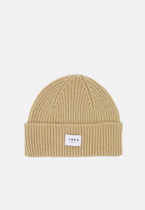 Knit beanie in light beige with a ribbed texture and a fold-over cuff featuring a white label with black text.