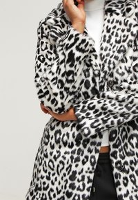 Persona vistiendo un abrigo de piel sintética de leopardo en blanco y negro, con un brazo cruzado y la otra mano tocándose la barbilla.