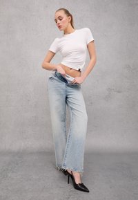 White cropped t-shirt, faded light blue wide-leg jeans with frayed hem, and black high-heeled shoes. Model poses with hands on jeans.